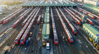 Multiple passenger trains parked on parallel tracks at a railway maintenance depot with storage buildings and rail yard infrastructure.