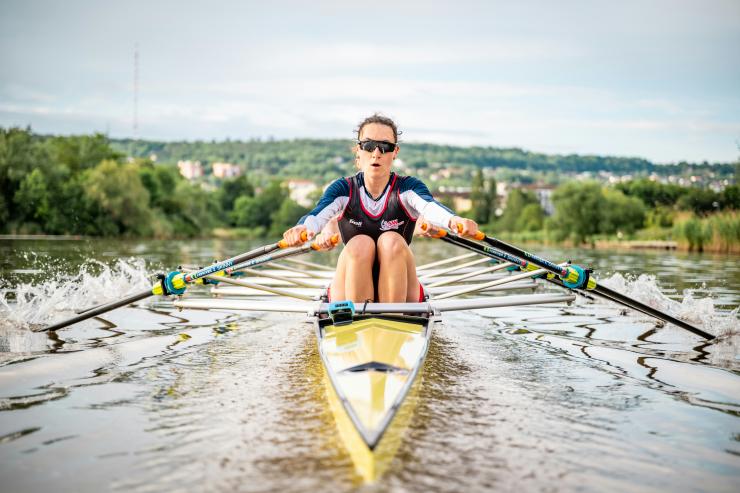 Front-facing view of a single sculler rowing on a calm river, wearing sunglasses and athletic gear, pulling two oars in sync as water splashes outward, with a green landscape and distant buildings in the background