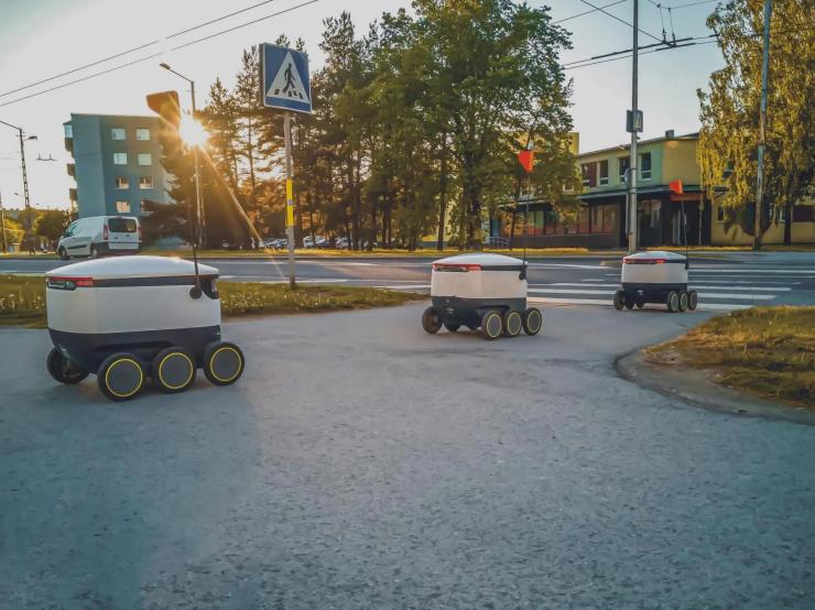 Three autonomous mobile delivery robots navigating an urban sidewalk near a pedestrian crossing