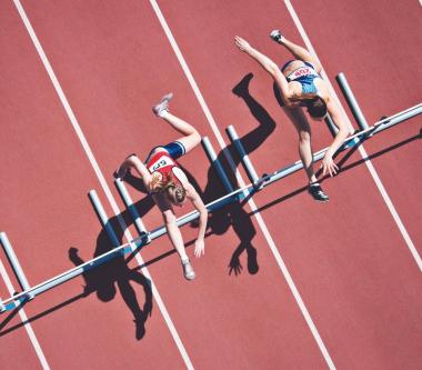 Two athletes jumping over hurdles on a running track, viewed from above